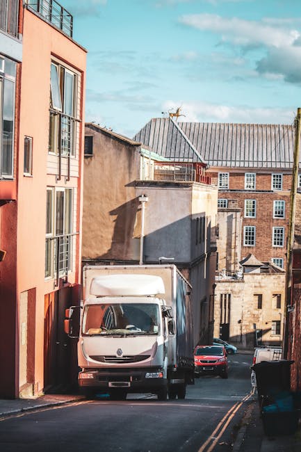 Image showing a residential street with a white moving van parked on the narrow roadway between multi-storey buildings, including a red-brick structure and a modern pink apartment block. The van is positioned close to a building entrance, with its rear doors visible. Several cardboard boxes and wrapped furniture items can be seen inside the van, indicating an ongoing home relocation process. The street surface is asphalt with yellow traffic lines, and a small black car is parked further down the street. A black waste bin is placed near the curb on the right side of the image. The sky is partly cloudy with blue patches, and shadows from the buildings suggest late morning or early afternoon sunlight. This scene reflects typical aspects of planning and executing furniture transport and packing and moving services, as provided by companies like Man and a Van Islington for tight access property moves, especially in urban areas.