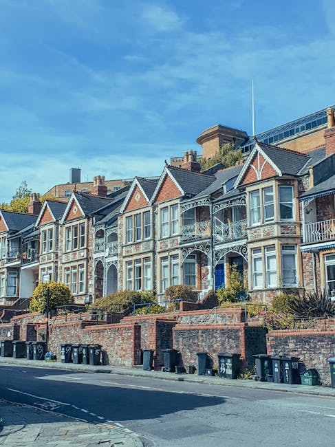 A row of Victorian terraced houses with ornate white ironwork balconies and decorative trim, situated on a slightly elevated street level with brick retaining walls and small front gardens. The houses have bay windows, pitched roofs with chimneys, and are built from brick with contrasting light-colored facades. In the foreground, there is a paved road with black wheelie bins lined up along the curb. The scene is captured during daylight with a bright blue sky overhead, and the setting appears to be in a residential area of Islington, suitable for home relocation or furniture transport services provided by Man and a Van Islington as part of their removals offerings.