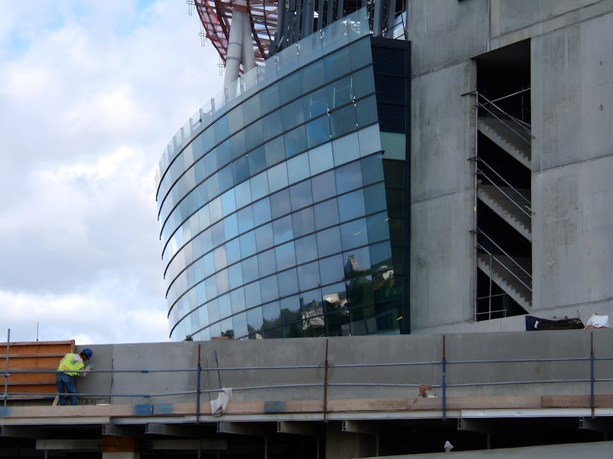 A construction worker wearing a high-visibility yellow jacket and a blue helmet is positioned on a partially completed concrete balcony, securing a long orange wooden formwork panel with ties. The worker is standing on the edge of the balcony next to a blue safety railing. In the background, a modern, curved glass building with reflective windows showcases the surrounding cityscape, while the sky above is partly cloudy. The construction site features an open staircase embedded within the concrete structure, with metal railings. The environment indicates an active building development area, with external construction materials and equipment visible. The image highlights aspects of building scaffolding and loading zones, relevant to house removals, home relocation, or furniture transport, which companies like Man and a Van Islington may be involved in during off-site packaging and transport activities.