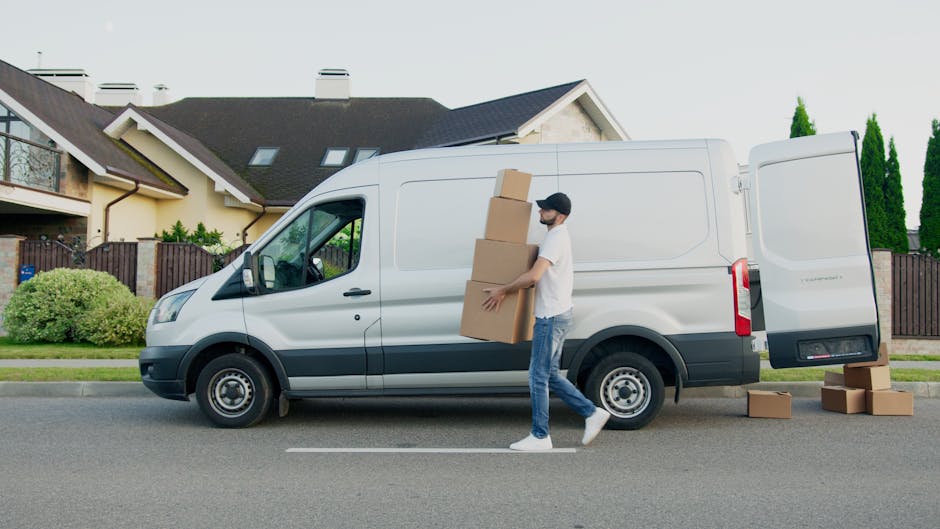 A man wearing a black cap, white t-shirt, and jeans is loading three medium-sized cardboard boxes into the side door of a white cargo van parked on a residential street. The van is positioned on the roadway, with its rear door open, revealing a partially visible interior suitable for removals and home relocation. Several other cardboard boxes are on the pavement next to the van, awaiting transportation or unloading. The background features a modern, multi-story house with a sloped roof, dormer windows, and a small front garden with green bushes and trees, situated behind a dark wooden fence. The sky appears overcast, and the scene depicts a typical furniture transport or packing and moving process conducted by a professional removal service like Man and a Van Islington, demonstrating the loading stage of a house move.