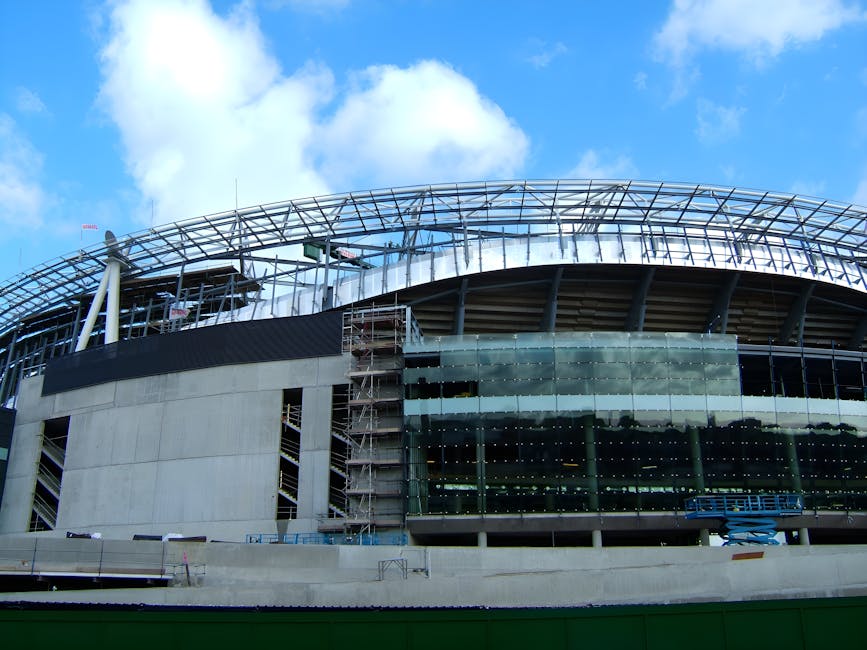 A modern building under construction with a curved glass facade and scaffolding on one side, showing an open roof structure supported by white steel beams, situated against a bright blue sky with scattered clouds. The lower part of the building features concrete walls and large tinted glass windows, while construction equipment such as a blue cherry picker and safety barriers are visible on the site. The scene depicts the building in the early stages of development, linked to ongoing construction and refurbishment activities that are typical in home relocation projects, where careful planning and logistics are essential. The exterior environment is well-lit with natural daylight, highlighting the urban setting for property improvements or commercial development, as managed by companies like Man and a Van Islington during comprehensive house removals and furniture transport processes.