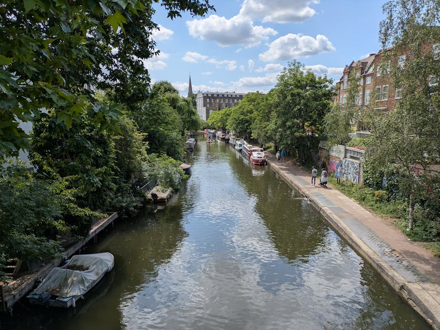 A wide view of a narrow canal in Islington during daytime, with calm water reflecting the partly cloudy sky above. On the left side, dense green trees with leafy branches extend over the water, with a small boat docked alongside the bank, covered by a light protective tarp. On the right side, a paved walkway runs parallel to the canal, with several parked cars and a few pedestrians walking or standing. The background features historic brick residential buildings with large windows and decorative facades, and a church steeple rises above the trees and structures. The scene depicts an urban riverside environment typical of a home relocation area, where the natural setting complements the nearby buildings and suggests a context for the moving services provided by Man and a Van Islington, involved in furniture transport and packing during house removals.