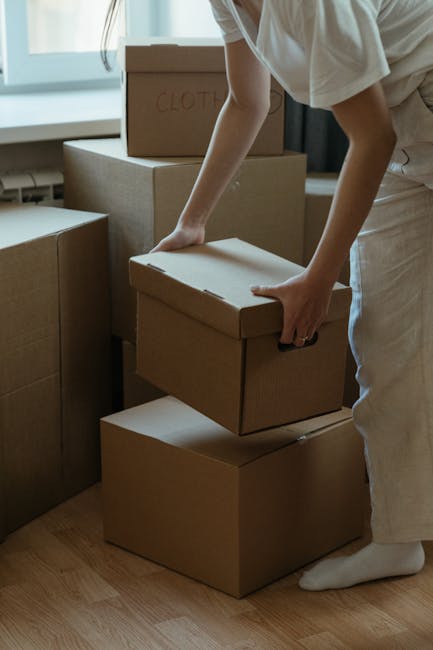 In the interior of a property, a person dressed in light clothing is seen placing a cardboard box onto a stack of similar boxes on a wooden floor, with natural light coming through a window behind the