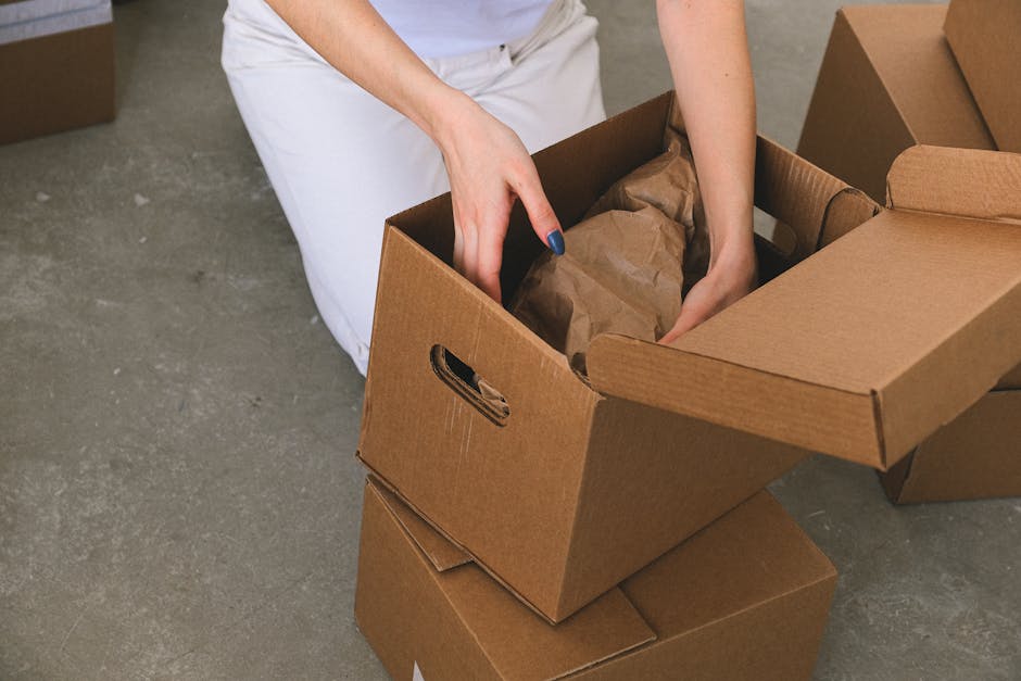 In the interior of a property, a person dressed in light clothing is seen placing a cardboard box onto a stack of similar boxes on a wooden floor, with natural light coming through a window behind the