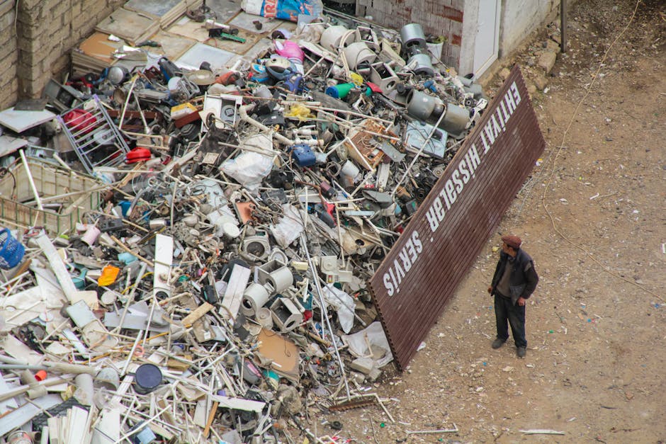 Two men are engaged in a house removal process involving waste disposal in an outdoor area. One man, dressed in a black jacket and yellow gloves, is lifting a cardboard box filled with plastic waste a