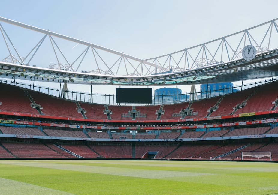 The image depicts the interior of a large, empty football stadium with a well-maintained green pitch in the foreground. Surrounding the pitch are multiple tiers of red and grey seating arranged in a semi-circular pattern, with some sections featuring banners displaying team names and sponsors. Above the seating area is a structural white roof supported by a network of metal trusses, which extends over part of the stands and includes a large digital scoreboard hanging centrally. The sky above is clear and bright, illuminating the empty stands and field. This environment illustrates the scale and design of a professional sports venue, which could be relevant in the context of moving or relocating sports equipment, stadium furniture, or similar large assets. The image is neutral and factual, focusing only on visual elements without additional context, suitable for discussing logistics involved in stadium or large-scale venue removals, as might be detailed in content about managing sports facility relocations or arrangements, like those related to Islington council removal permits.