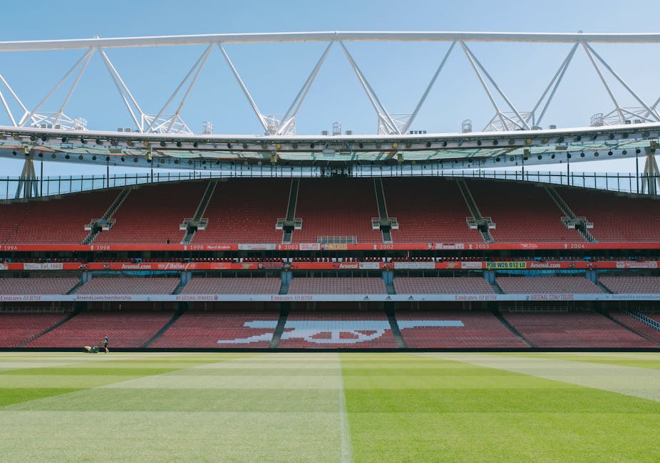 Inside a large sports stadium with a well-maintained green football pitch in the foreground, rows of red stadium seats extend from the field upwards. The seats are mostly empty, with some white markings forming a pattern on the central section. Above, a modern white roof structure supported by steel trusses spans across the upper part of the stadium, casting partial shadows over the seating area. The sky visible through the open sides of the roof is bright and clear, indicating daytime. This image, associated with house removals and relocation services, captures the spacious environment where moving logistics like furniture transport or packing might be coordinated before or after a home relocation, as referenced on the [PAGE_TITLE] page of Man and a Van Islington.