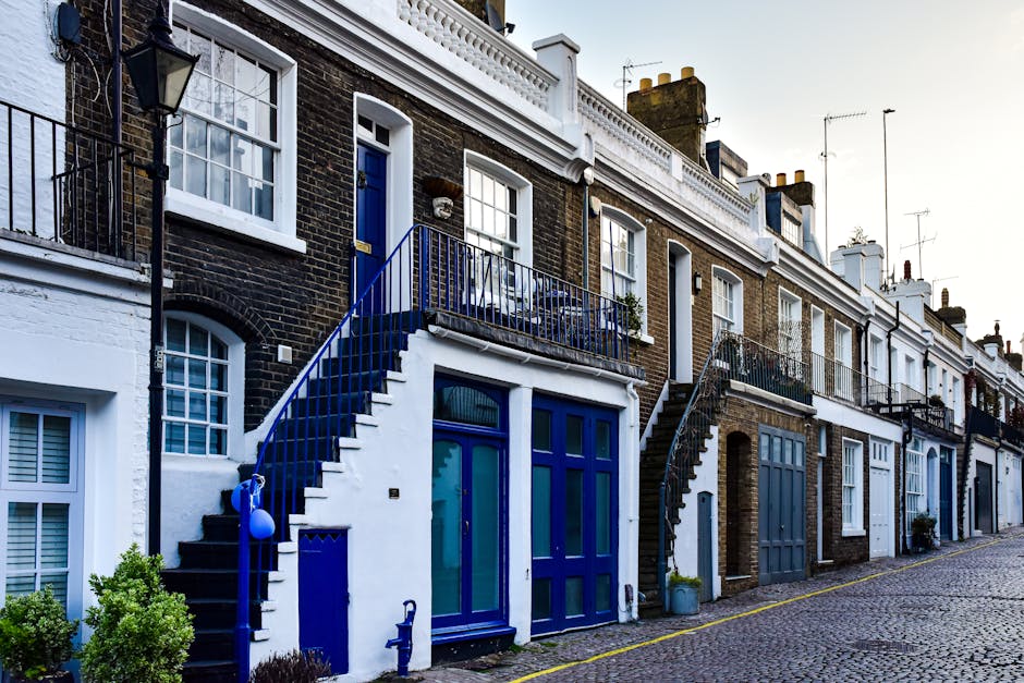 A row of Victorian-style terraced houses on a cobblestone street in Islington, featuring brick facades with white accents around windows and doors. Each property has a small external staircase with a metal railing leading up to a second-floor entrance, painted predominantly in shades of blue. Some staircases have black treads and railings, while others are blue, with potted plants and small decorative items visible on some balconies. The ground-floor entrances have large glass doors and windows, also framed in blue, with a variety of shrubs and potted plants placed outside. The street is lined with a yellow line, and the lighting suggests early morning or late afternoon. This setting provides a suitable backdrop for house removals and furniture transport, with an emphasis on the external environment and typical urban residential features, indicative of a home relocation scenario supported by companies like Man and a Van Islington.