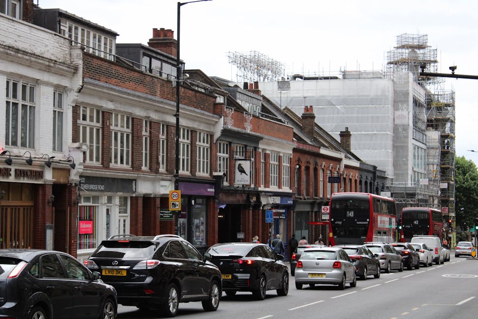 A street scene in Upper Street, Islington, showing a row of brick and sash-window terraced buildings with retail shops at ground level. In front, several parked cars line the street, including a black Mercedes, a white vehicle, and a silver car, while a double-decker bus marked with route number 343 is visible arriving or departing. On the pavement, a few pedestrians walk past the buildings, some approaching or leaving a shop entrance. Towards the right side of the image, there is ongoing building renovation with scaffolding and white protective sheeting covering a multi-storey structure, indicating construction work. The scene is captured during daylight hours under overcast weather, with natural lighting illuminating the urban environment. This setting reflects a typical street where home relocation or furniture transport might involve logistical planning by professional removal services such as Man and a Van Islington, especially given the presence of diverse vehicles and ongoing construction related to property improvements or conversions.