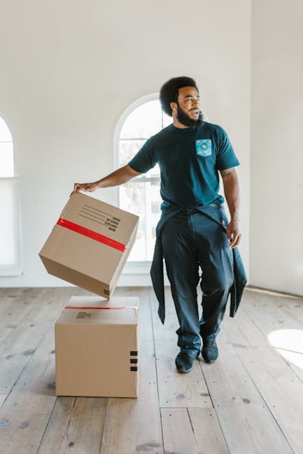 A man with dark curly hair and a beard, dressed in a navy blue t-shirt and matching trousers, is inside a bright, minimally furnished room with large arched windows allowing natural light to fill the space. He is holding a cardboard box wrapped with red tape in his right hand, about to place it on top of another similar box on the wooden floor. The boxes are made of brown corrugated cardboard, and the room features light-colored wooden flooring and plain white walls. In the background, an open doorway and the windows are visible, indicating an ongoing home relocation process. The scene depicts the packing and loading stage typical of professional removals, with the man preparing boxes for transport, possibly as part of a furniture transport or packing and moving service offered by Man and a Van Islington.