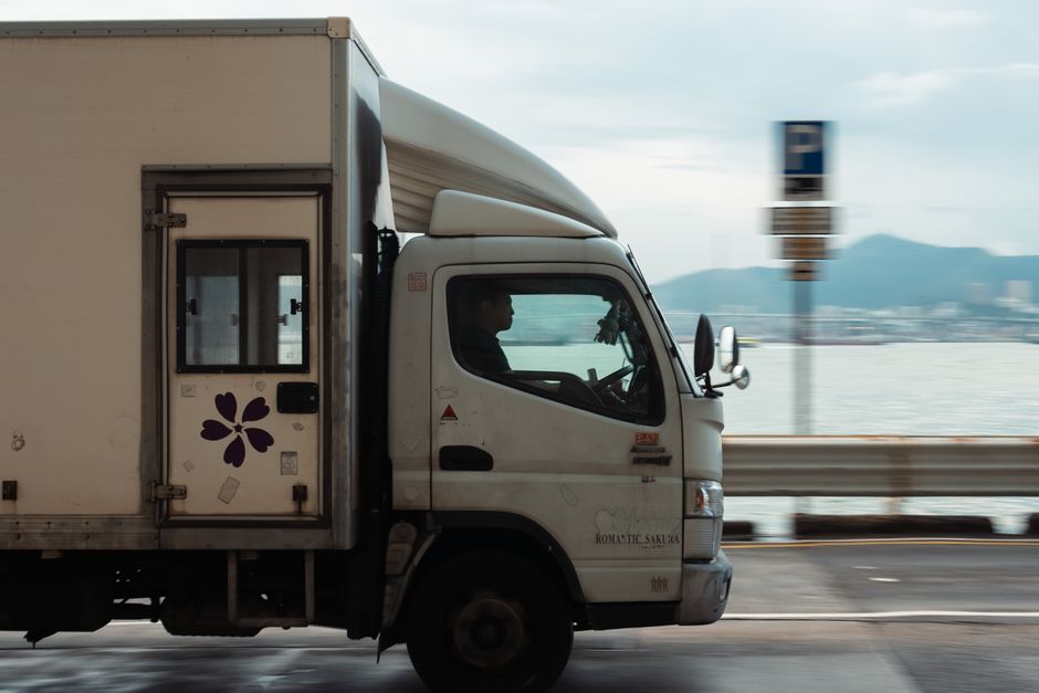 Side view of a white moving truck with decorative purple flower decals parked near a waterfront, with a driver visible inside the cab. The truck is positioned on a road with a metal guardrail separating it from the water, which extends into a body of water with mountains in the background. The truck's cargo area is closed, and the vehicle is likely involved in house removals or home relocation services provided by Man and a Van Islington. The scene suggests a loading or transit phase in the furniture transport process, with natural daylight illuminating the environment and a parking sign in the background, indicating an urban or suburban area suitable for residential moves.