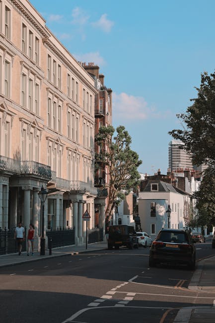 A street scene in Islington with a large, multi-story residential building featuring light-colored brickwork, decorative window details, and small balconies on the left side. Several white and dark-colored cars are parked along the curb and a few are driving on the road. In the background, there are trees with green foliage and a modern high-rise building under a blue sky with some clouds. An individual is walking on the pavement near the building, and a man and woman are standing nearby. The scene captures the typical urban environment associated with house removals and relocation activities in London, with a focus on the architectural details and street traffic. Man and a Van Islington specializes in local removals, including packing and furniture transport, and this image visually aligns with their services involving home relocation and loading processes.
