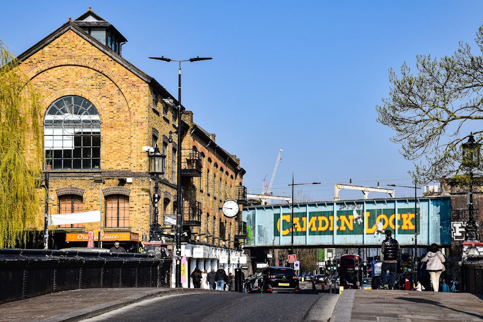 A wide street scene featuring a brick building with large arched windows and a clock mounted on the facade, situated alongside a railway bridge painted in teal with the words 'CAMDEN LOCK' in yellow signage. The street is populated with pedestrians walking on the pavement, some carrying bags, and several parked cars lining the street. Overhead, streetlights and lampposts are visible, along with a high railway track with supporting ironwork. In the background, cranes and construction equipment suggest nearby development. The scene is well-lit with clear blue sky overhead, depicting an urban environment potentially related to house removals or furniture transport activities, as could be coordinated by companies like Man and a Van Islington during live moving or home relocation processes.